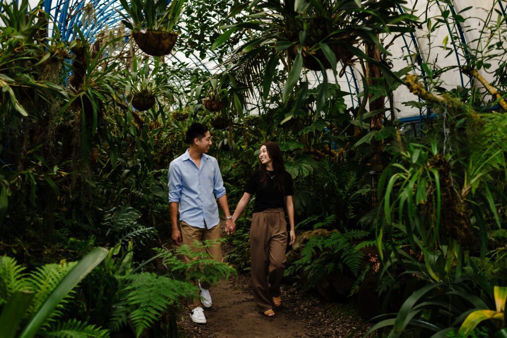 Novios caminando de la mano a través del exuberante orquideario del Jardín Botánico de Quito. Una captura llena de vida y colores reales que documenta la conexión natural de la pareja, por Viviana Calaón.