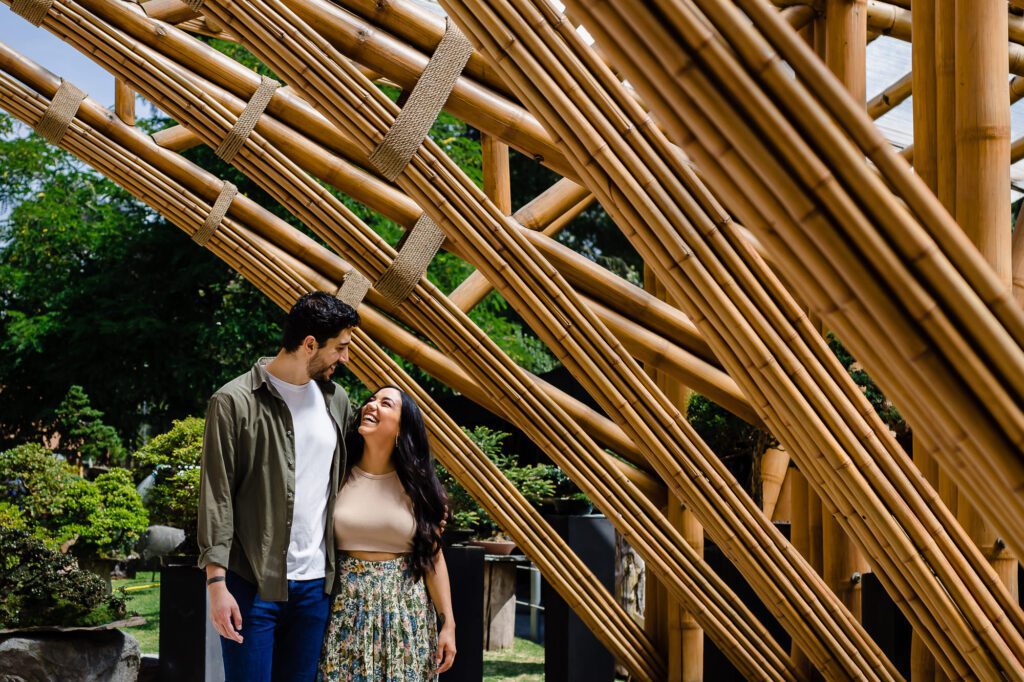 Momento espontáneo de una pareja compartiendo risas bajo una estructura de bambú en el Jardín Botánico de Quito. Fotografía de pedida de mano enfocada en emociones reales y sin poses forzadas por Che Vivi.