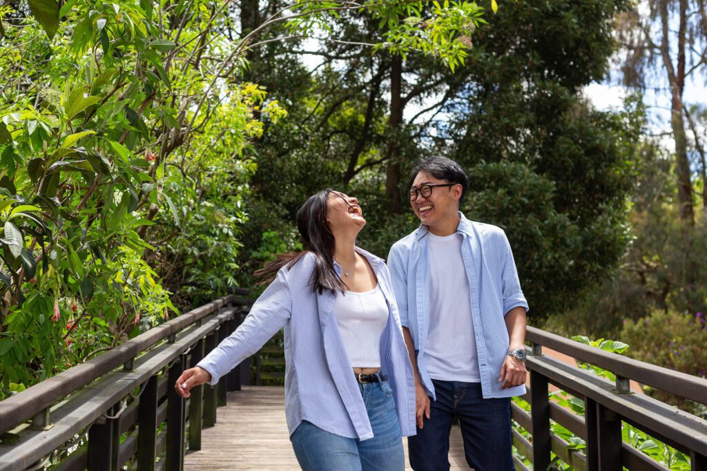 Explosión de alegría y risas naturales de una pareja cruzando un puente de madera en Jardín Botánico de Quito. En Che Vivi nos enfocamos en que recordés tus emociones, no las poses, capturando tu esencia más auténtica.