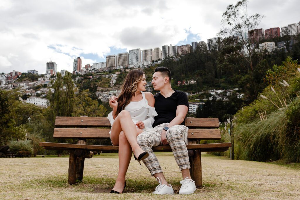 Pareja sentada de forma relajada en un banco de madera con la ciudad de Quito de fondo. Fotografía que refleja paz y felicidad real sin presiones ni poses rígidas.