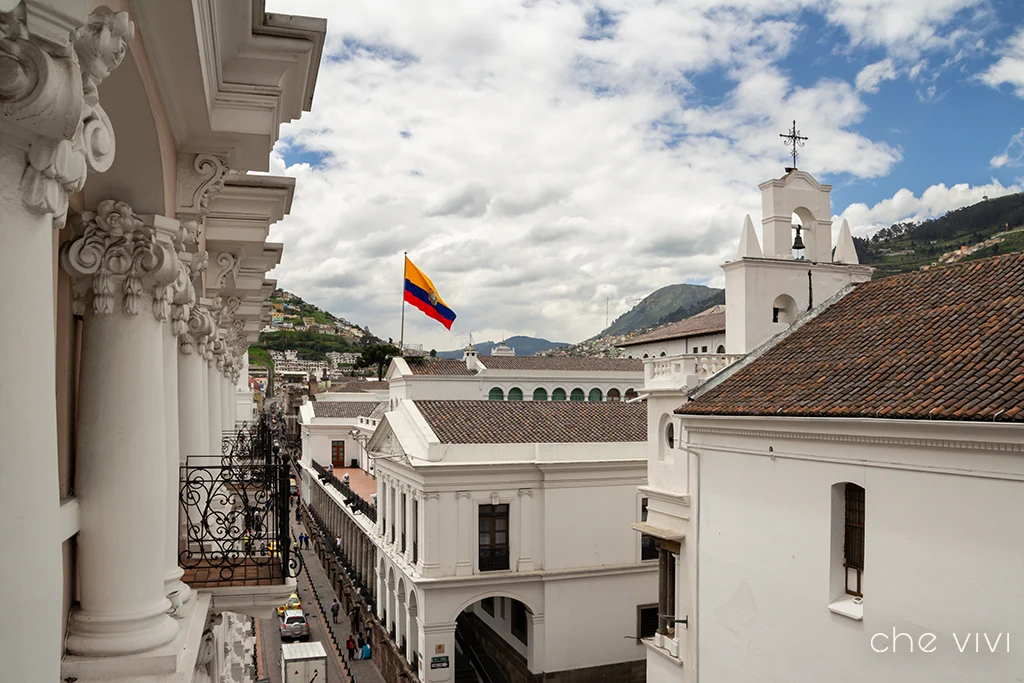 Perspectiva desde un balcón del Hotel Plaza Grande hacia la arquitectura colonial y tejados del Centro Histórico de Quito, con la bandera de Ecuador flameando. El entorno patrimonial perfecto para una propuesta de matrimonio.
