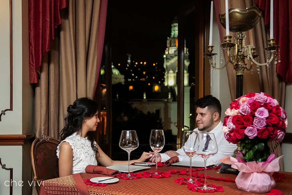 Pareja compartiendo una cena romántica a la luz de las velas y tomándose de la mano junto a la ventana del Hotel Plaza Grande en Quito. Las iglesias iluminadas del Centro Histórico de fondo.